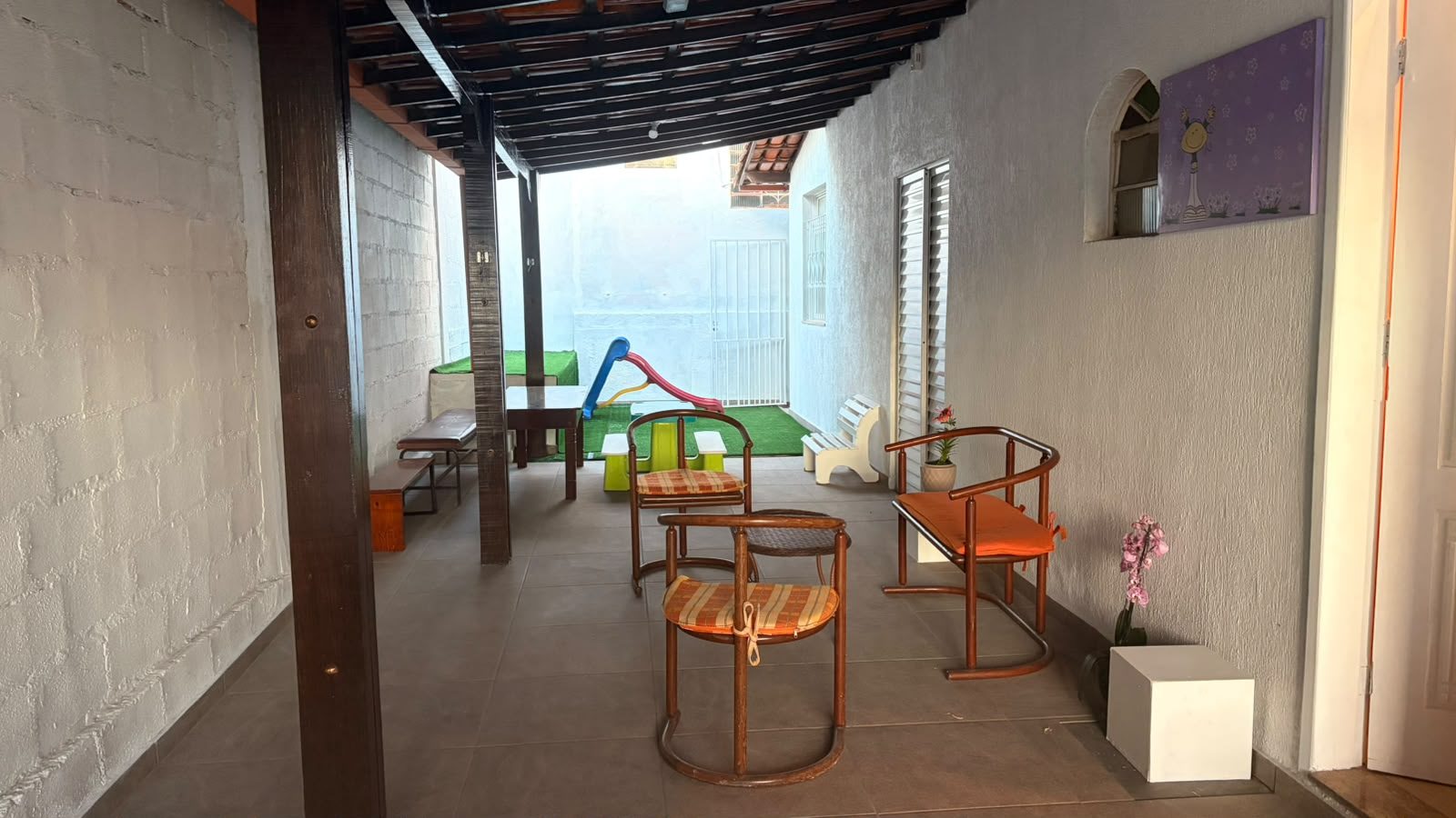 Área de convivência coberta com cadeiras e, ao fundo, espaço infantil com escorregador.