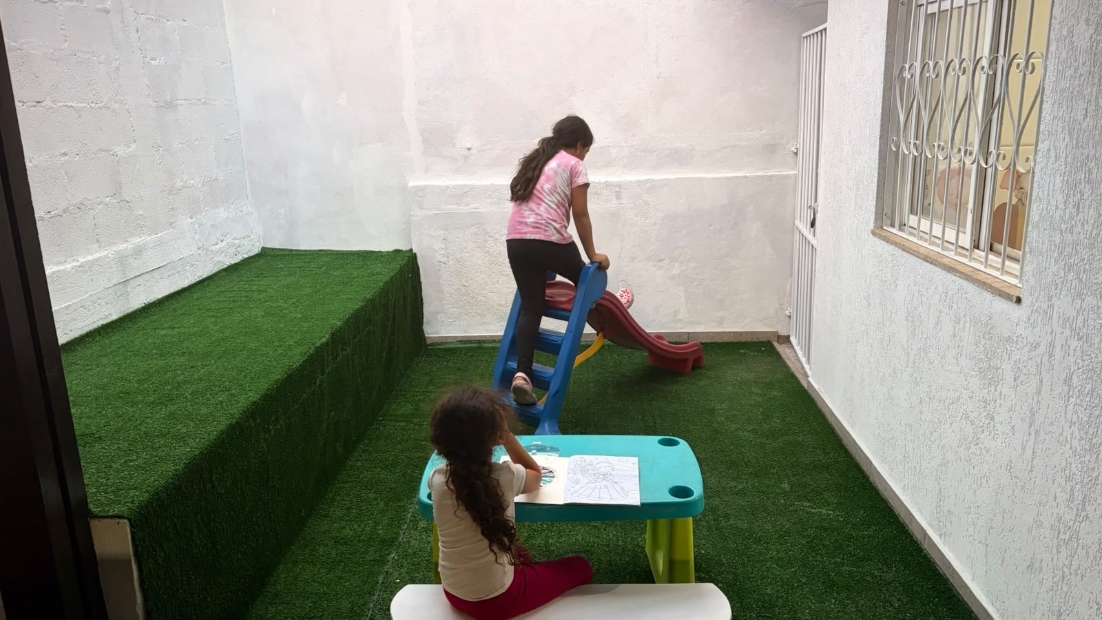 Área externa infantil com grama sintética, mesa para atividades e escorregador.