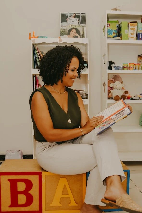 Dra. Jennifer Barbosa sentada sobre blocos coloridos, lendo um livro infantil.