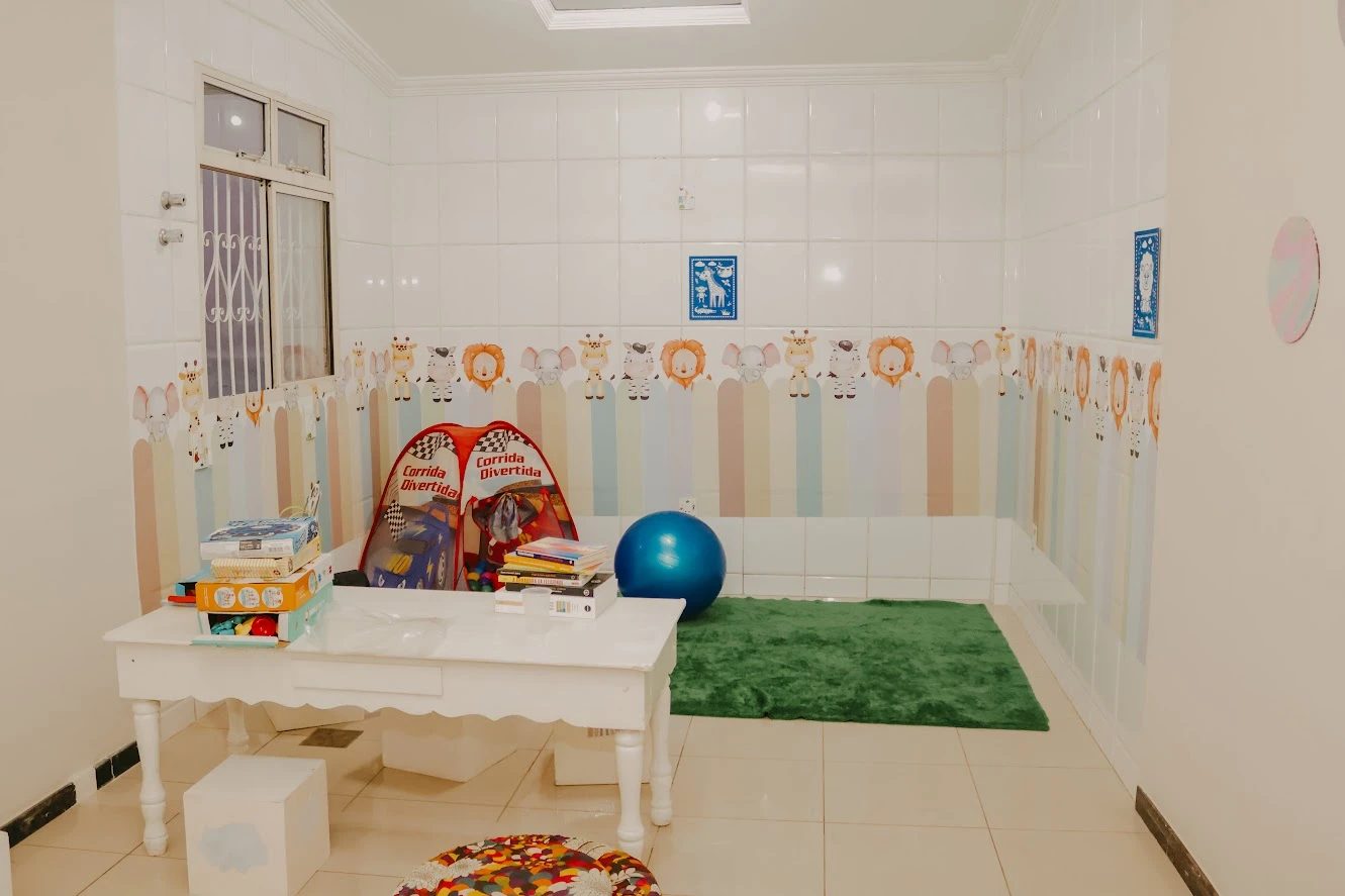 Sala infantil com parede decorada com animais, tenda de brinquedo, bola de exercício, tapete verde e mesa com jogos e livros.