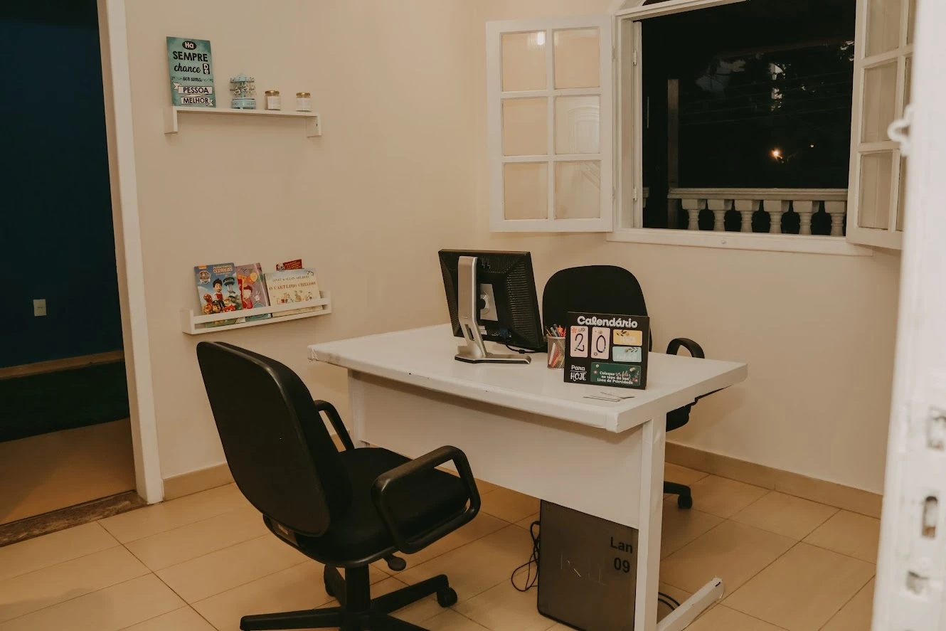 Secretaria com mesa, computador, cadeiras e prateleiras com livros infantis.